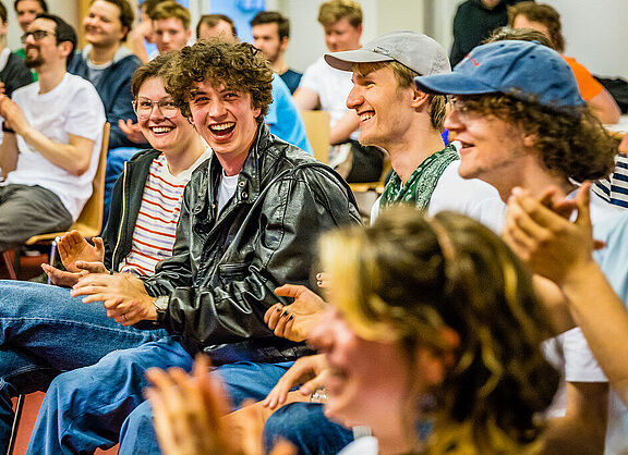 Viele Menschen sitzen in einem Raum, klatschen und lachen. Die Menschen sehen jung aus. In der Bildmitte sitzt ein junger Mann in Lederjacke und mit lockigen Haaren. 